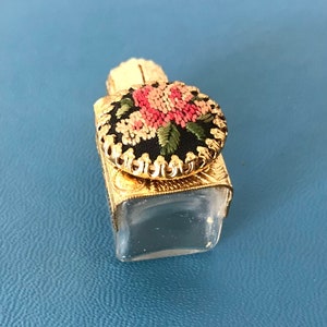 Op de afbeelding: Een klein, doorzichtig glazen flesje met een gouden metalen dop. De dop is versierd met een zwarte en witte geborduurde bloem met roze en groene accenten.