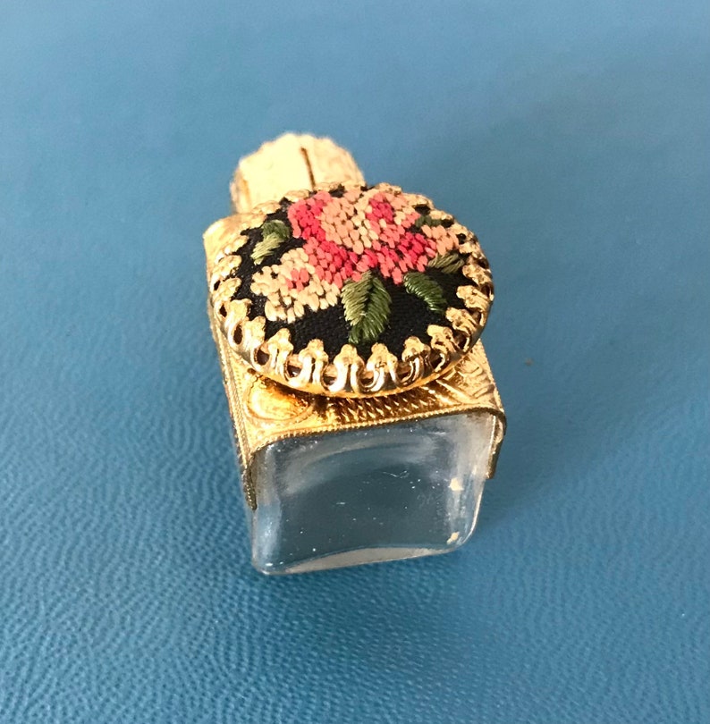 Op de afbeelding: Een klein, doorzichtig glazen flesje met een gouden metalen dop. De dop is versierd met een zwarte en witte geborduurde bloem met roze en groene accenten.