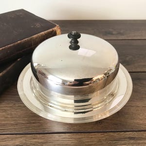 May include: A silver butter dish with a domed lid and a black knob. The dish sits on a round base with a ridged design. Two antique books with dark brown covers are in the background, adding a vintage feel.