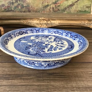 May include: A round ceramic pedestal plate featuring a blue and white willow pattern. The plate has a raised circular base and a wide rim decorated with intricate geometric and floral motifs, while the center depicts a traditional landscape scene.
