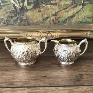 May include: A silver sugar bowl and creamer set with detailed floral patterns. The sugar bowl has two handles, and the creamer has a spout. The items are displayed on a wooden surface, with a framed painting visible in the background.