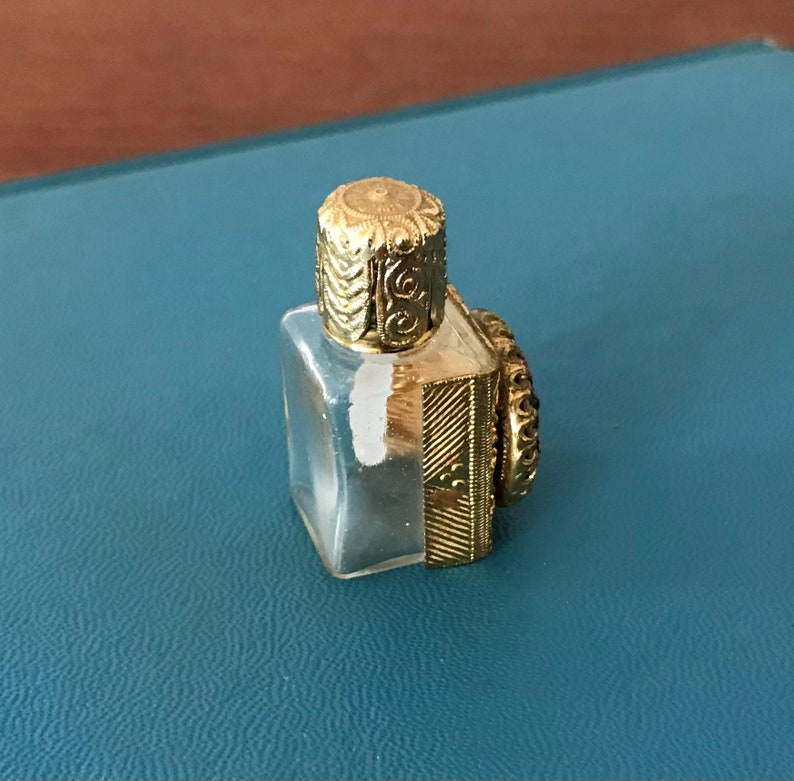 Op de afbeelding: Een klein, doorzichtig glazen flesje met een gouden dop en ingewikkelde details. Het flesje heeft een vierkante vorm en een decoratief ontwerp aan de zijkant.