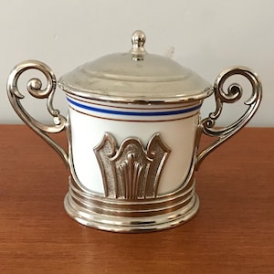 May include: A silver sugar bowl with a white porcelain base and a blue and gold trim. The bowl has a lid and two ornate handles.