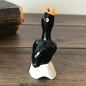 Peut inclure: Figurine en céramique représentant un oiseau noir avec une base blanche et un bec orange. L'oiseau a une finition brillante et un seul œil blanc. La figurine est sur une surface en bois.