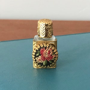Op de afbeelding: Een klein, goudkleurig glazen flesje met een bloemenmotief. Het flesje heeft een helder glazen lichaam en een gouden dop. Het bloemenmotief is geborduurd met roze en witte bloemen op een zwarte achtergrond.