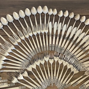 May include: An assortment of silver-colored silverware, including spoons, forks, and knives, arranged in a circular pattern on a wooden surface. The silverware has decorative detailing on the handles, suggesting a formal dining set.