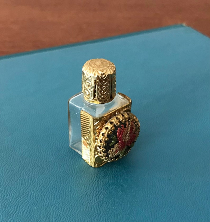 Op de afbeelding: Een klein, helder glazen flesje met een gouden dop en een decoratief bloemmotief aan de voorkant. Het flesje staat op een blauw oppervlak.