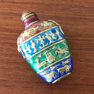 May include: A small, oval-shaped, multi-colored, enamel bottle with a gold lid. The bottle is decorated with gold painted animals and designs. The bottle is on a wooden surface.