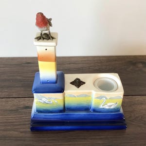 May include: A ceramic decorative piece with a bird on top, a small cup, and a swan design. The piece is painted in shades of blue, yellow, and white.