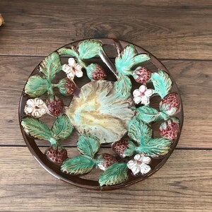 May include: A round ceramic trivet featuring a raised relief design of strawberries, green leaves, and white blossoms. The center has a mottled beige and brown glaze, while the outer border is a dark brown. The piece has an openwork structure.