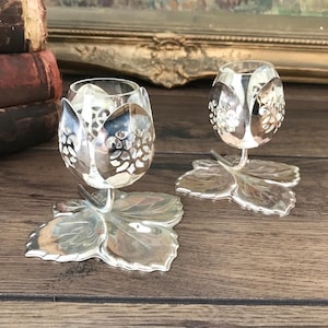 May include: Two silver-toned metal candle holders, each featuring a base shaped like a large leaf and a top designed as a tulip flower with intricate cut-out patterns. The tulip-shaped cups are supported by a curved stem rising from the leaf base.