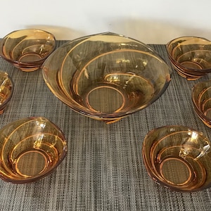 Op de afbeelding: Een set van zes amberkleurige glazen kommen, waaronder een grote serveerschaal en vijf kleinere kommen. De kommen hebben een draaiende, textuur.