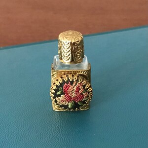 Op de afbeelding: Een klein, helder glazen flesje met een gouden metalen dop en een bloemenmotief aan de voorkant. Het flesje is versierd met een gouden metalen frame en een bloemenmotief.