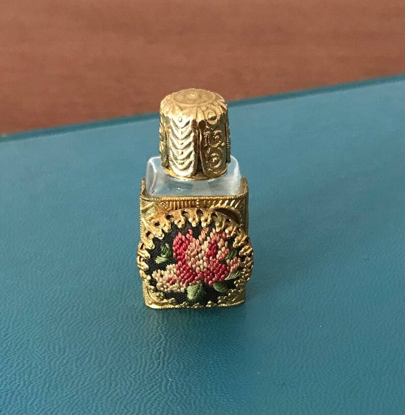 Op de afbeelding: Een klein, helder glazen flesje met een gouden metalen dop en een bloemenmotief aan de voorkant. Het flesje is versierd met een gouden metalen frame en een bloemenmotief.