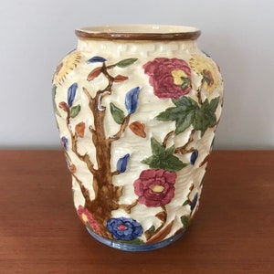 May include: A cream-colored ceramic vase with a raised floral design. The vase features a tree with brown branches, green leaves, and pink, blue, and yellow flowers.