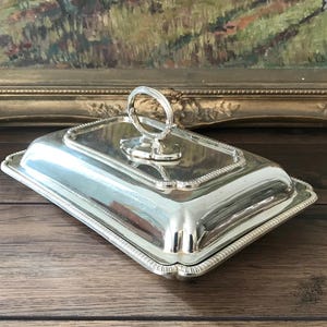 Op de afbeelding: Een rechthoekige, verzilverde serveerschaal met deksel en handvat. De schaal heeft een decoratieve, parelrand en staat op een houten oppervlak. Op de achtergrond staat een ingelijst schilderij.