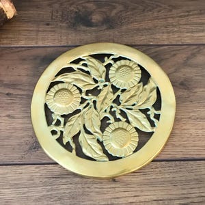 May include: A circular brass trivet featuring an openwork design of three sunflower blossoms surrounded by detailed leaves and stems. The metal surface has a polished gold-toned finish with a smooth outer rim and intricate cutouts throughout.