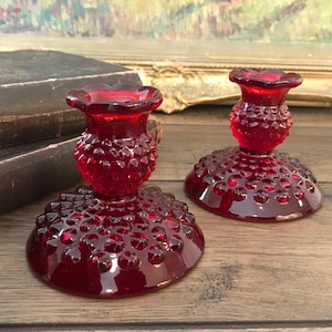May include: Two ruby red glass candle holders with a hobnail design. The candle holders have a round base and a flared top. The candle holders are sitting on a wooden surface.