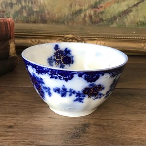 May include: A white porcelain bowl featuring a deep blue floral pattern around the exterior and interior rim. The design includes intricate dark blue botanical motifs with subtle gold-colored accents and a scalloped edge along the top rim.