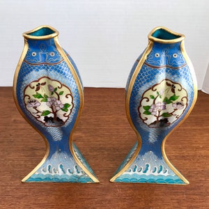 May include: A pair of blue and gold cloisonn&eacute; vases shaped like fish with floral designs. The vases are decorated with white, pink, and green flowers and leaves.