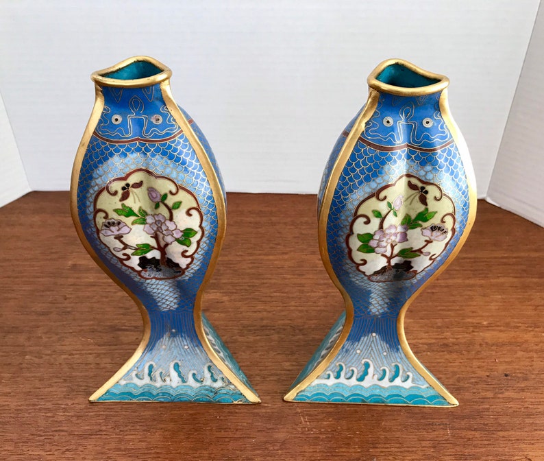 May include: A pair of blue and gold cloisonn&eacute; vases shaped like fish with floral designs. The vases are decorated with white, pink, and green flowers and leaves.