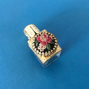 Op de afbeelding: Een klein, doorzichtig glazen flesje met een gouden metalen dop. De dop is versierd met een zwart en gouden bloemenmotief. Het flesje is waarschijnlijk voor parfum of etherische oli&euml;n.