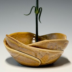 May include: A single pink carnation in a small, handcrafted, ceramic vase. The vase is a warm, earthy brown with a unique, layered design. The flower's stem is dark green, and the petals are a soft, delicate pink.