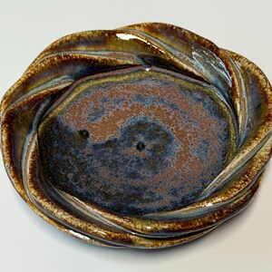 May include: A handmade ceramic bowl with a unique, twisted rim design. The bowl features a blend of colors, including brown, blue, and gold, creating a textured, glazed finish. The center of the bowl is a darker shade of blue and brown.