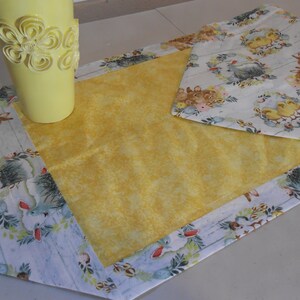 Può includere: Un runner da tavolo giallo con un bordo bianco e un motivo floreale con coniglietti e pulcini. Il runner è su una superficie bianca con un vaso giallo con un design floreale.