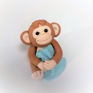 May include: A brown monkey figurine holding a light blue blanket. The monkey has a smiling face and is sitting on a white background.