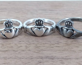 May include: Three silver Claddagh rings with a heart, crown, and hands design. The rings are on a wooden surface.