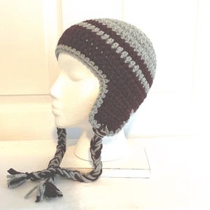 May include: A handmade crochet hat in shades of gray and burgundy, featuring ear flaps and braided ties with tassels. The hat is displayed on a white mannequin head, showcasing its textured stitch and color-blocked design. A cozy winter accessory.