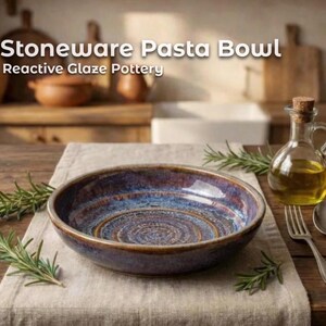 Könnte beinhalten: Steinzeug-Pastaschüssel mit reaktiver Glasurkeramik. Die Schüssel hat einen dunkelbraunen Rand und einen blau-braunen Innenraum mit einem Spiraldesign. Eine Glasflasche mit Öl, eine Gabel und ein Löffel stehen auf dem Tisch.