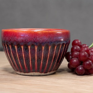 May include: A ceramic bowl with a glossy, gradient finish, transitioning from red to dark purple. Vertical grooves create a textured pattern on the bowl's exterior. A bunch of red grapes rests beside the bowl on a wooden surface.