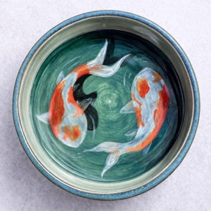 Puede incluir: Un cuenco de cerámica con borde azul e interior verde. Dos peces koi, con marcas naranjas y blancas, están pintados dentro del cuenco, nadando en círculo. El cuenco está sobre una superficie blanca.