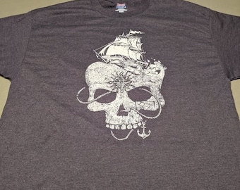 Puede incluir: Una camiseta gris oscuro con un gráfico blanco de una calavera con un barco navegando fuera de su cabeza. La calavera está rodeada de una cuerda y un ancla.