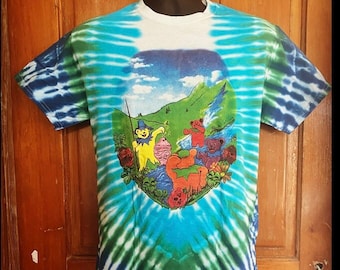 Puede incluir: Una camiseta tie-dye con un patrón de espiral azul, verde y blanco. La parte delantera de la camiseta presenta un gráfico de los osos danzantes de los Grateful Dead en un entorno de bosque.