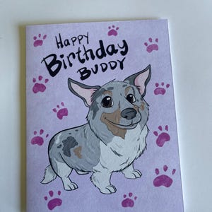 May include: A birthday card featuring a cartoon dog illustration. The card reads "Happy Birthday Buddy" in black lettering. The dog is depicted in shades of grey, white, and brown, with pink paw prints scattered around.