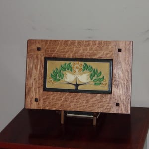May include: A rectangular ceramic tile featuring a relief design of two white birds perched in a tree with green leaves and yellow berries, set within a thick, light brown oak wood frame with four small square dark inlays at the corners.