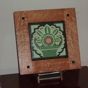 May include: Square wooden frame with a decorative tile insert. The tile features a green and pink floral design within a black border. The frame is made of light brown wood with dark square accents at each corner.