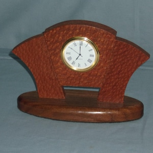 May include: A wooden desk clock with a gold-tone clock face and Roman numerals. The clock is set in a reddish-brown, fan-shaped wooden frame, and sits on a dark brown oval base. The clock face has black hands and a white background.