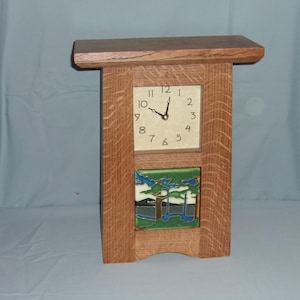 May include: A wooden Arts and Crafts style clock with a square face and a decorative tile. The clock face has black numerals and hands. The tile features a landscape scene with blue, green, and white colors.