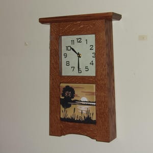 May include: A rectangular wooden wall clock with a square clock face and a landscape tile. The clock face has black numerals and hands. The landscape tile depicts a tree, water, and a sunset. The clock is mounted on a wall.