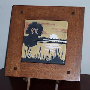 May include: A square ceramic tile featuring a relief landscape scene of a tree, cattails, and a setting sun, framed in dark brown wood with four small square corner accents. The tile displays muted tones of tan, black, and white in a nature motif.