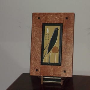 May include: A wooden framed art piece featuring a black bird and floral design on a yellow background. The frame is a light brown color with square cutouts in each corner. The artwork is displayed on a dark brown surface.