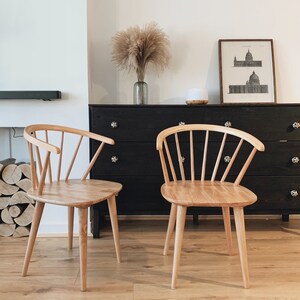 Peut inclure: Deux chaises en bois clair avec un dossier &agrave; fuseaux et un si&egrave;ge arrondi. Les chaises sont devant une commode noire avec une estampe encadr&eacute;e au-dessus.