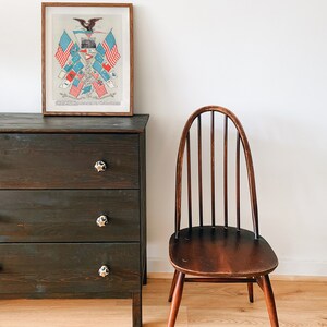 May include: A dark wood dresser with three drawers and black knobs. A wooden chair with a curved back and a wooden seat sits to the right of the dresser. A framed print of an eagle with American flags hangs on the wall above the dresser.