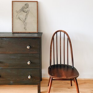 May include: A dark wood dresser with three drawers and black knobs. A wooden chair with a curved back and spindles sits to the right of the dresser. A framed drawing of a ballerina hangs on the wall above the dresser.