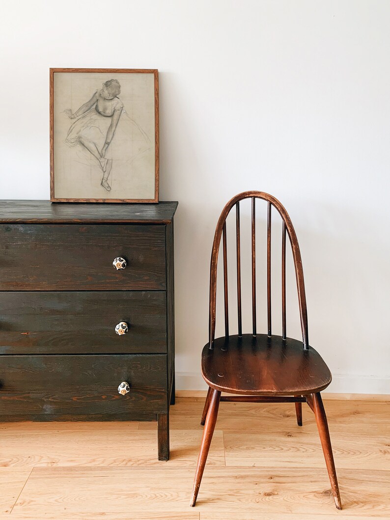 May include: A dark wood dresser with three drawers and black knobs. A wooden chair with a curved back and spindles sits to the right of the dresser. A framed drawing of a ballerina hangs on the wall above the dresser.