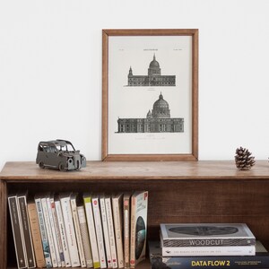 Peut inclure: Une estampe en noir et blanc encadr&eacute;e d'un dessin architectural d'un b&acirc;timent avec un d&ocirc;me. L'estampe est sur une &eacute;tag&egrave;re en bois avec une petite voiture jouet, des livres et des pommes de pin.